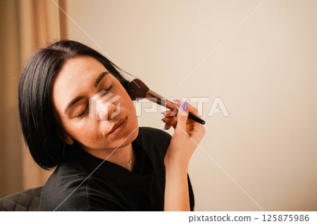 A close-up of a woman applying makeup captures her gentle technique with a fluffy brush. Her closed eyes convey concentration. The minimalist background enhances the focus on her routine. 125875986