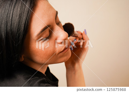 Her closed eyes convey concentration. The minimalist background enhances the focus on her routine. A close-up of a woman applying makeup captures her gentle technique with a fluffy brush. 125875988
