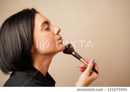 The minimalist background enhances the focus on her routine. A close-up of a woman applying makeup captures her gentle technique with a fluffy brush. Her closed eyes convey concentration. 125875994
