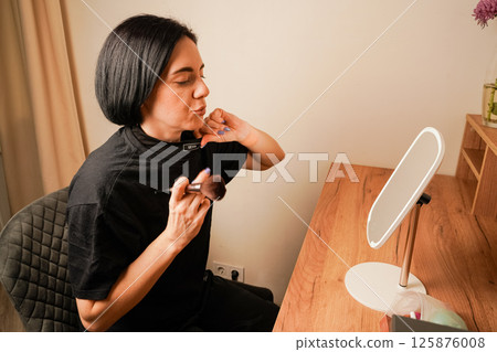 Her delicate features are reflected in the table mirror. Her dark hair completes her look. Focusing on her makeup, the woman uses a soft brush to enhance her complexion. Her delicate features are reflected in the table mirror. Her dark hair completes her look. Focusing on her makeup, the woman uses a soft brush to enhance her complexion. 125876008
