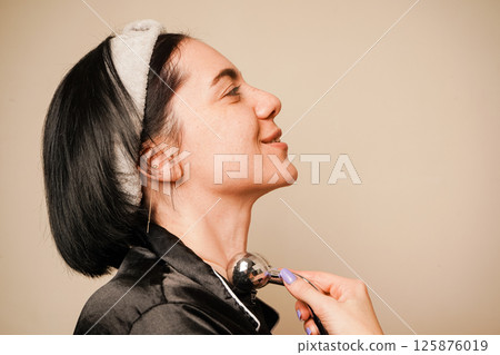 A woman enjoys a relaxing facial massage with a metal roller. Her headband holds her hair back, revealing her smooth complexion. The soft lighting emphasizes her natural beauty. A woman enjoys a relaxing facial massage with a metal roller. Her headband holds her hair back, revealing her smooth complexion. The soft lighting emphasizes her natural beauty. 125876019