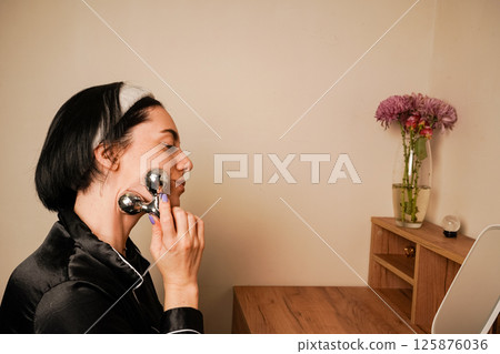 Her expression reflects self-care. A woman uses a delicate metal roller to massage her jawline for a soothing skincare treatment. A girl in black pajamas sits at a table in front of a mirror. 125876036