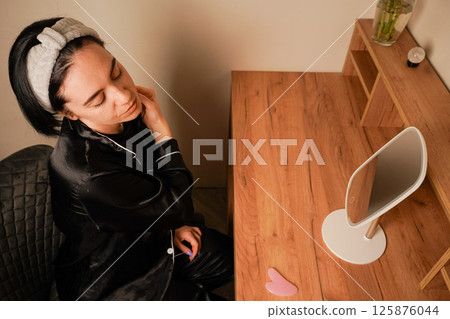 Her expression reflects self-care. A woman uses a delicate metal roller to massage her jawline for a soothing skincare treatment. A girl in black pajamas sits at a table in front of a mirror. 125876044