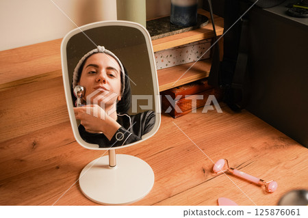 Young woman doing anti-aging facial massage with a metal roller. Reflection of brunette during massage in table mirror. Girl with a headband holding her hair. Young woman doing anti-aging facial massage with a metal roller. Reflection of brunette during massage in table mirror. Girl with a headband holding her hair. 125876061