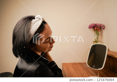 Reflection of brunette during massage in table mirror. Girl with a headband holding her hair. Young woman doing anti-aging facial massage with a metal roller. 125876064