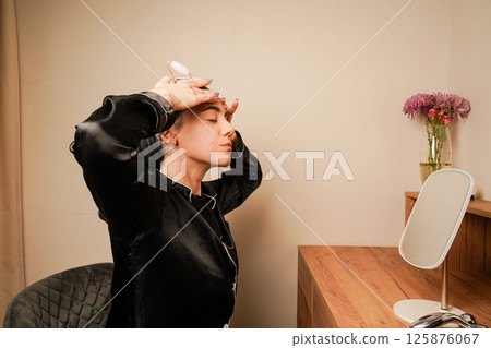 Woman in black pajamas and with a headband doing beauty procedures at home. Girl doing facial massage with her hands. Caring for aging skin. Applying cream to face. 125876067