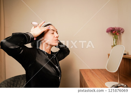 Girl doing facial massage with her hands. Caring for aging skin. Applying cream to face. Woman in black pajamas and with a headband doing beauty procedures at home. Girl doing facial massage with her hands. Caring for aging skin. Applying cream to face. Woman in black pajamas and with a headband doing beauty procedures at home. 125876069