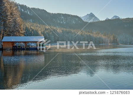 Serene Winter Lakeside Sunrise with Snow-Covered Boathouse Reflecting in Calm Waters: Majestic Mountain Landscape Bathed in Golden Light Serene Winter Lakeside Sunrise with Snow-Covered Boathouse Reflecting in Calm Waters: Majestic Mountain Landscape Bathed in Golden Light 125876083