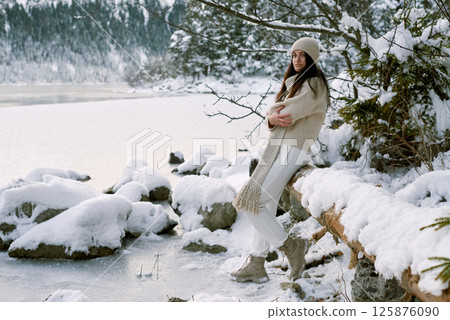Serene winter portrait of woman in beige knitwear standing on snow-covered log by frozen lake surrounded by snowy evergreen forest Serene winter portrait of woman in beige knitwear standing on snow-covered log by frozen lake surrounded by snowy evergreen forest 125876090