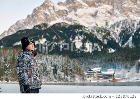 Young boy standing on a frozen lake in a snowy winter landscape with evergreen forest and majestic mountains in the background on a clear cold day 125876122