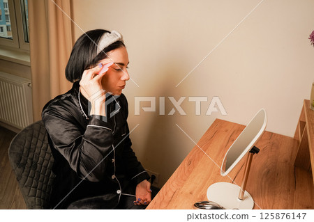 A woman in black pajamas uses a gua sha tool to massage her face and neck. The headband holds her hair back, emphasizing her glowing skin. A peaceful at-home beauty routine is depicted. 125876147