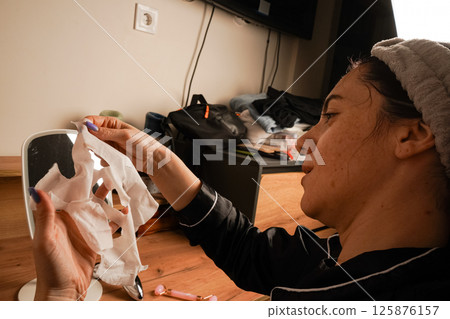 Wearing a headband and black silk pajamas, a woman takes care of her skin with a moisturizing mask. A small mirror on the table reflects her radiant skin. A girl's reflection in a table mirror. 125876157