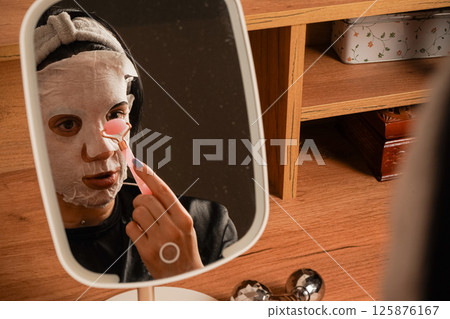 A small mirror on the table reflects her radiant skin. A girl's reflection in a table mirror. Wearing a headband and black silk pajamas, a woman takes care of her skin with a moisturizing mask. A small mirror on the table reflects her radiant skin. A girl's reflection in a table mirror. Wearing a headband and black silk pajamas, a woman takes care of her skin with a moisturizing mask. 125876167