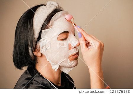 A young woman is using a hydrating mask at her dressing table. She is wearing elegant black pajamas and a headband. A small mirror adds charm to the scene. 125876188