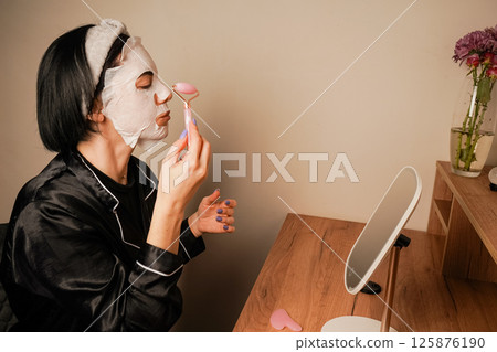 A small mirror adds charm to the scene. A young woman is using a hydrating mask at her dressing table. She is wearing elegant black pajamas and a headband. 125876190