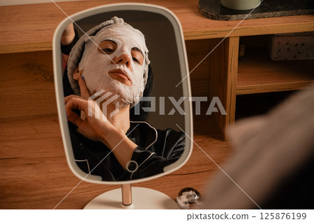 Black pajamas and a neat headband complete the scene. A small mirror reflects her calm expression. Sitting at a makeup table, a woman relaxes with a hydrating sheet mask. Black pajamas and a neat headband complete the scene. A small mirror reflects her calm expression. Sitting at a makeup table, a woman relaxes with a hydrating sheet mask. 125876199