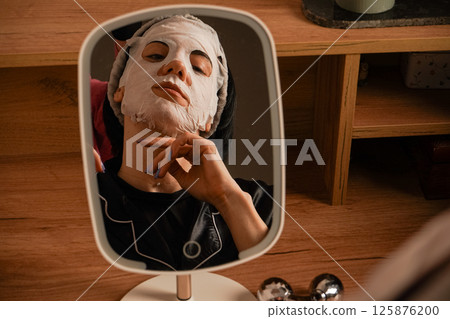 A small mirror reflects her calm expression. Sitting at a makeup table, a woman relaxes with a hydrating sheet mask. Black pajamas and a neat headband complete the scene. A small mirror reflects her calm expression. Sitting at a makeup table, a woman relaxes with a hydrating sheet mask. Black pajamas and a neat headband complete the scene. 125876200
