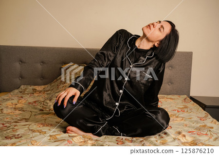 Her short black hair frames her face softly. A young woman wakes up and stretches in her black silk pajamas. She makes gentle hand and head movements to energize her body. 125876210