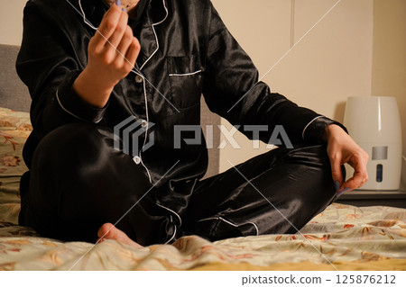 View of the girl's legs. Close-up of the details of the pajamas. Close-up of a young woman sitting in silk black pajamas, starting her day with morning stretching. View of the girl's legs. Close-up of the details of the pajamas. Close-up of a young woman sitting in silk black pajamas, starting her day with morning stretching. 125876212