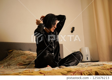 The morning begins with a young woman stretching gracefully in black silk pajamas. She moves her head and arms to awaken her senses after sleep. Morning ritual with vigorous exercise. 125876213