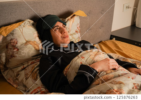 Dressed in silky black pajamas, she is surrounded by beautiful floral-patterned bedding. The setting radiates calm and relaxation. In a tranquil moment, a woman sleeps with a green mask covering her 125876263