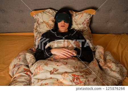Peacefully lying in bed, a woman enjoys her rest with a silk eye mask. Her black pajamas and soft floral bedding create a serene atmosphere. Her relaxed posture highlights a restful sleep. Peacefully lying in bed, a woman enjoys her rest with a silk eye mask. Her black pajamas and soft floral bedding create a serene atmosphere. Her relaxed posture highlights a restful sleep. 125876266