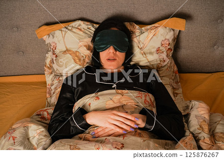 Her black pajamas and soft floral bedding create a serene atmosphere. Her relaxed posture highlights a restful sleep. Peacefully lying in bed, a woman enjoys her rest with a silk eye mask. Her black pajamas and soft floral bedding create a serene atmosphere. Her relaxed posture highlights a restful sleep. Peacefully lying in bed, a woman enjoys her rest with a silk eye mask. 125876267