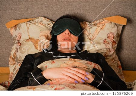 Her relaxed posture highlights a restful sleep. Peacefully lying in bed, a woman enjoys her rest with a silk eye mask. Her black pajamas and soft floral bedding create a serene atmosphere. 125876269
