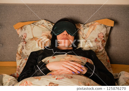 Her black pajamas and soft floral bedding create a serene atmosphere. Her relaxed posture highlights a restful sleep. Peacefully lying in bed, a woman enjoys her rest with a silk eye mask. 125876270