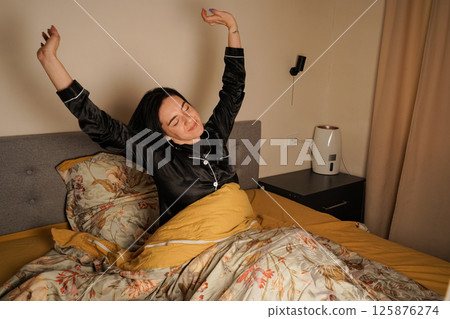 Wearing a black silk pajama set, she greets the morning with a smile. The warm tones of the room create a cozy atmosphere. Wearing a black silk pajama set, she greets the morning with a smile. The warm tones of the room create a cozy atmosphere. 125876274