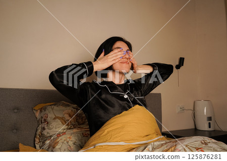 Dressed in elegant black silk pajamas, she begins her day with a cozy and relaxed moment. The floral bedding adds warmth to the scene. A young woman rubs her eyes gently after waking up in bed. 125876281
