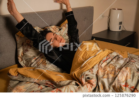 The warm tones of the room create a cozy atmosphere. A young woman stretches her arms joyfully while sitting in bed. Wearing a black silk pajama set, she greets the morning with a smile. 125876290