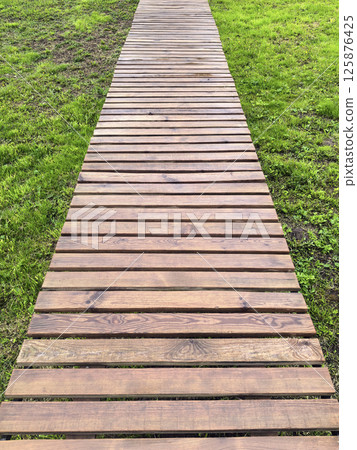 A path made of wooden planks on a background of green grass. Wooden trail in landscape design. Natural materials in the landscape of the park and garden. Footpath and walking trail A path made of wooden planks on a background of green grass. Wooden trail in landscape design. Natural materials in the landscape of the park and garden. Footpath and walking trail 125876425