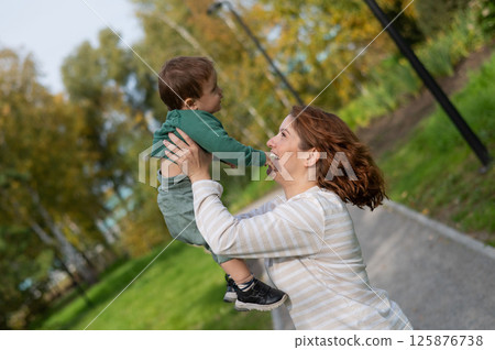 Caucasian woman holds her one-year-old son in her arms while walking in the park in autumn. Caucasian woman holds her one-year-old son in her arms while walking in the park in autumn. 125876738