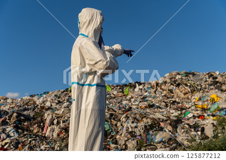 An environmental worker in protective clothing inspects a large landfill, pointing towards areas of concern amidst various waste An environmental worker in protective clothing inspects a large landfill, pointing towards areas of concern amidst various waste 125877212