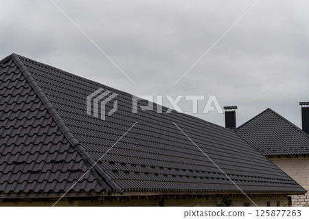 Dark metal roofs cover houses in a neighborhood as overhead clouds hint at possible rain. The scene captures a typical architectural design Dark metal roofs cover houses in a neighborhood as overhead clouds hint at possible rain. The scene captures a typical architectural design 125877263
