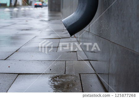 Water drips from a drain pipe onto slick pavement, showing the aftermath of rain in an urban environment 125877268