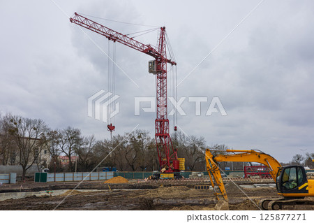A large red crane stands at a construction site with heavy machinery working amid overcast weather, indicating ongoing building activity A large red crane stands at a construction site with heavy machinery working amid overcast weather, indicating ongoing building activity 125877271