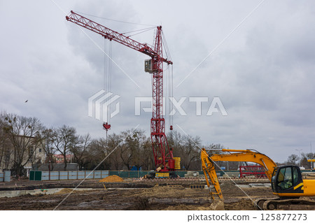 Heavy machinery operates at a construction site featuring a prominent red crane against a gray, cloudy sky, with trees in the background Heavy machinery operates at a construction site featuring a prominent red crane against a gray, cloudy sky, with trees in the background 125877273