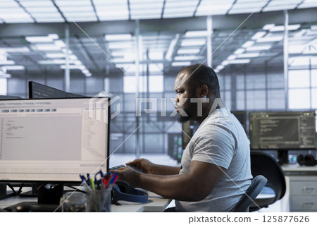 Technician at desk writing code and doing checkup on server room providing computing resources for different workloads. African american man investigating data center mainframes using computer 125877626