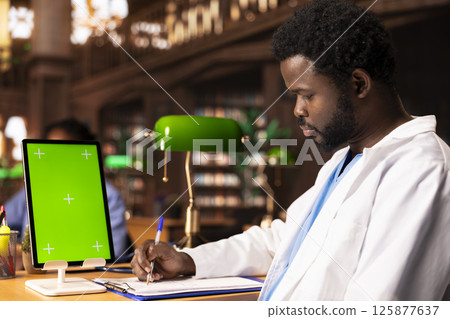African American med student reading a healthcare book next to green screen, focusing on medical practice methods under green lamps in a classic library. Commitment to education. 125877637