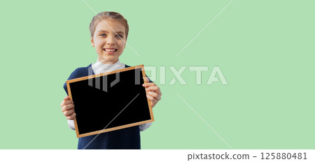 Banner happy 8 year old girl holds black mockup chalk board on green background, copy space 125880481