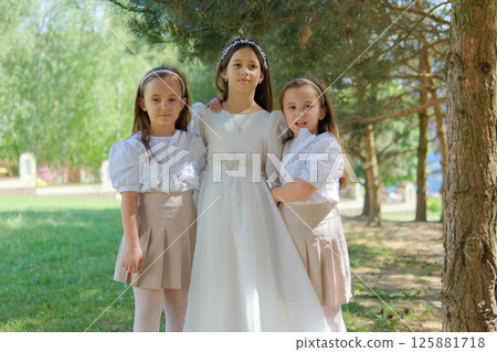 A little lady in a long ivory dress stands at the center of attention. Two younger girls gently hug the older child, emphasizing the family bond. A little lady in a long ivory dress stands at the center of attention. Two younger girls gently hug the older child, emphasizing the family bond. 125881718
