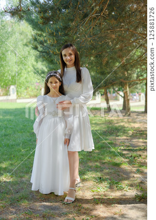A girl on the eve of her First Communion stands next to a young mother in elegant attire. A mother in a white dress hugs the child, offering warmth and support. 125881726
