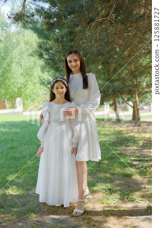 A girl on the eve of her First Communion stands next to a young mother in elegant attire. A mother in a white dress hugs the child, offering warmth and support. 125881727