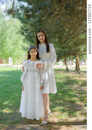 A little girl in a creamy long dress stands among the greenery of pine trees. A mother gently embraces the girl, creating a touching scene of unity A little girl in a creamy long dress stands among the greenery of pine trees. A mother gently embraces the girl, creating a touching scene of unity 125881728