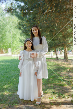 A little girl in a creamy long dress stands among the greenery of pine trees. A mother gently embraces the girl, creating a touching scene of unity 125881729