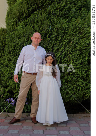 A girl in a long ivory dress stands beside her strong father. The father gently holds the child's hand, emphasizing the importance of the moment. 125881752