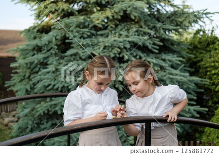 Two schoolgirls stand side by side, focused on the tiny creature. Dressed in identical light outfits, the children look exceptionally neat 125881772