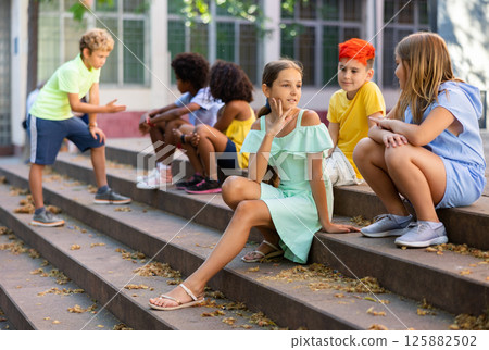 Cheerful preteen girls and boys sitting on steps outdoors and chatting Cheerful preteen girls and boys sitting on steps outdoors and chatting 125882502